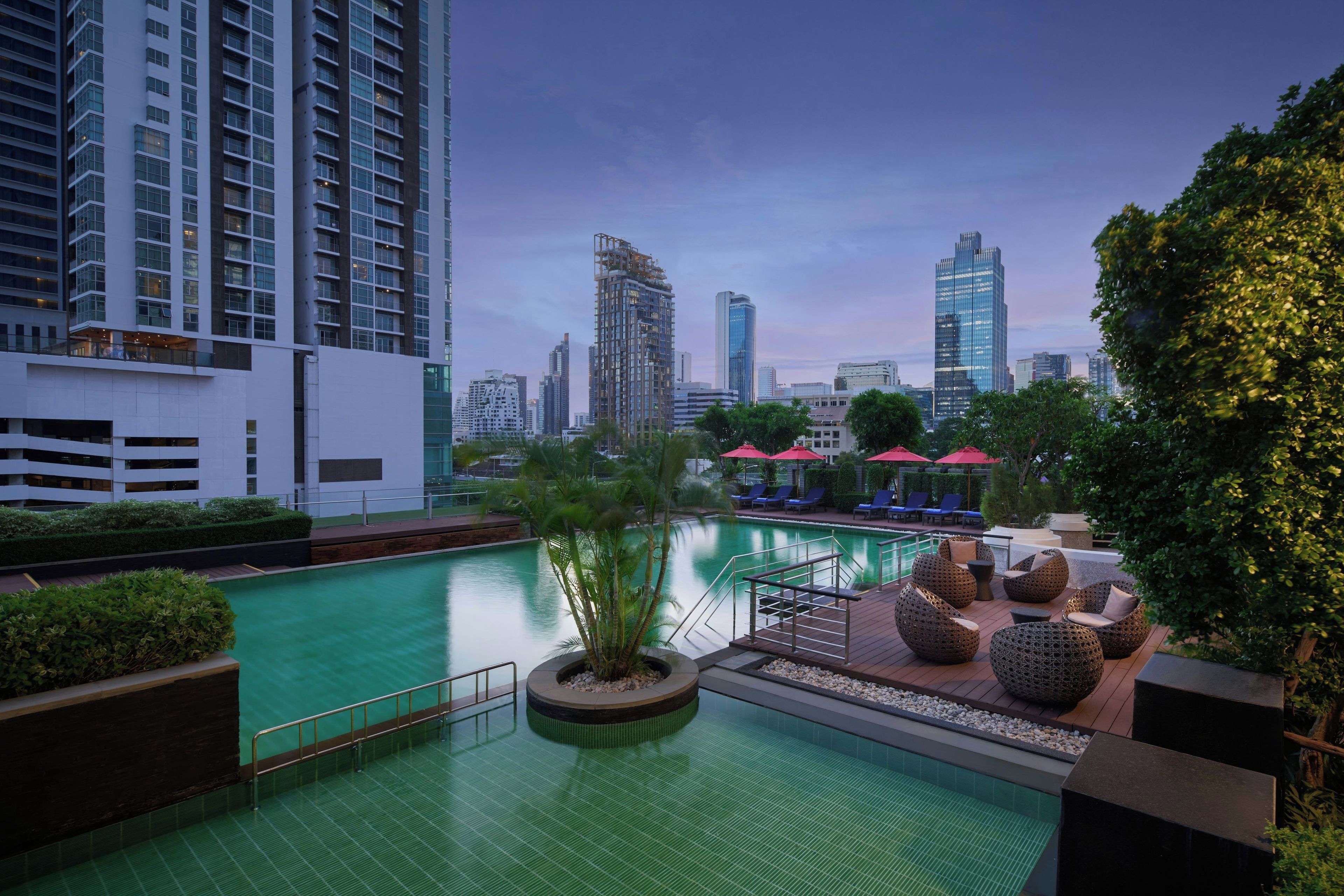 A rooftop pool with a view of a city skyline, surrounded by greenery and lounge chairs.