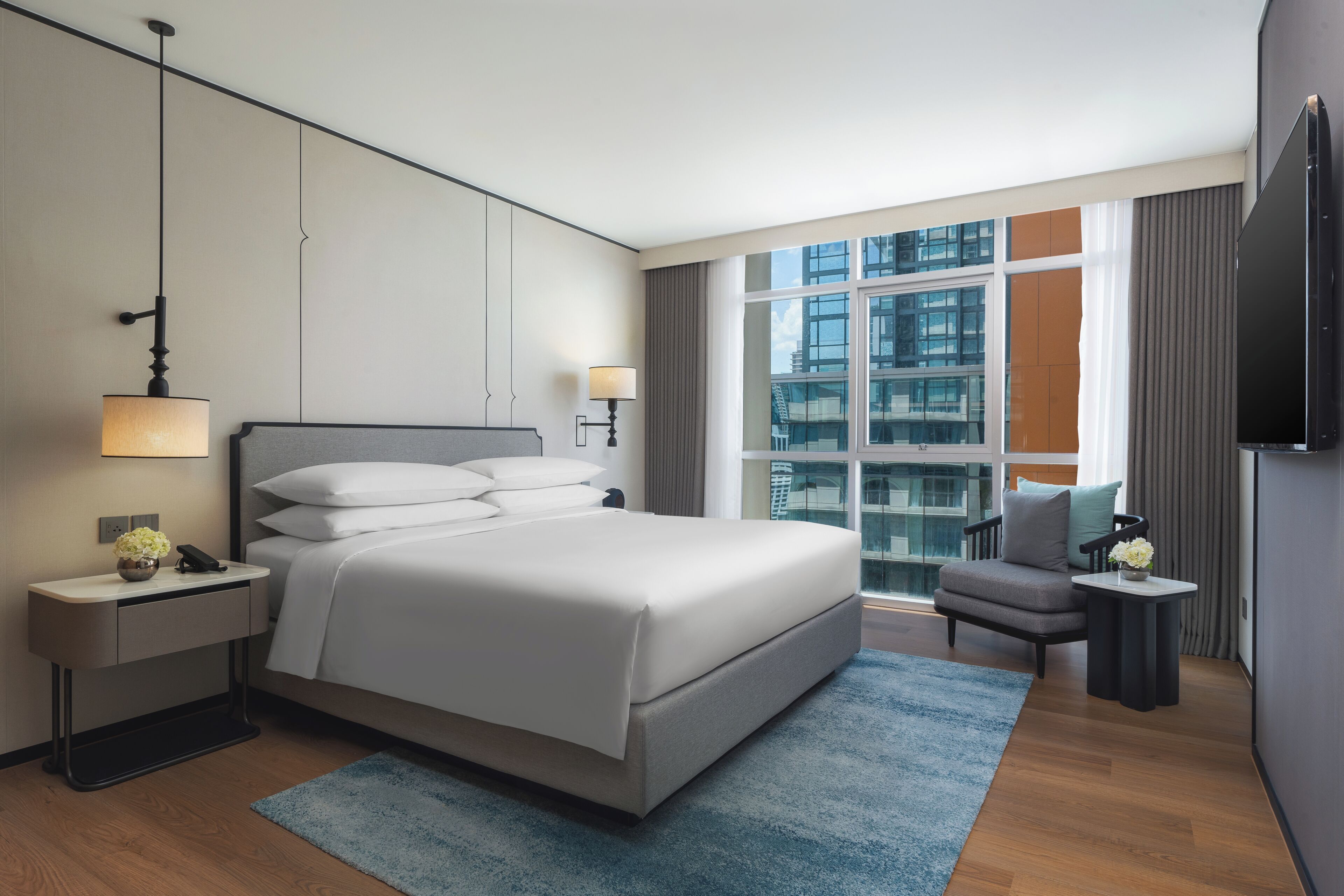 Habitación de hotel moderna con una cama grande, mesitas de noche, un escritorio, una silla, televisión y vistas a la ciudad.