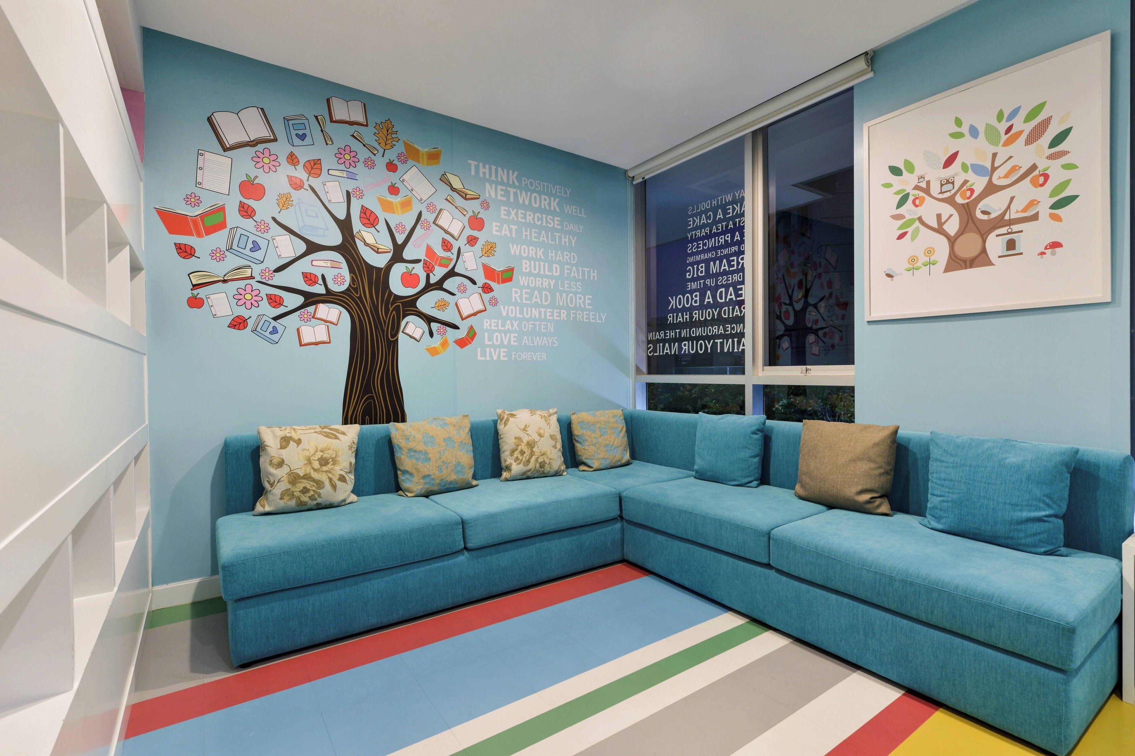 A modern living room with a blue sectional sofa, colorful striped rug, and a wall mural featuring a tree with books and positive phrases.