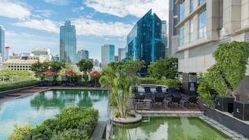 A pool area with lounge chairs and umbrellas, surrounded by greenery and modern buildings in the background.