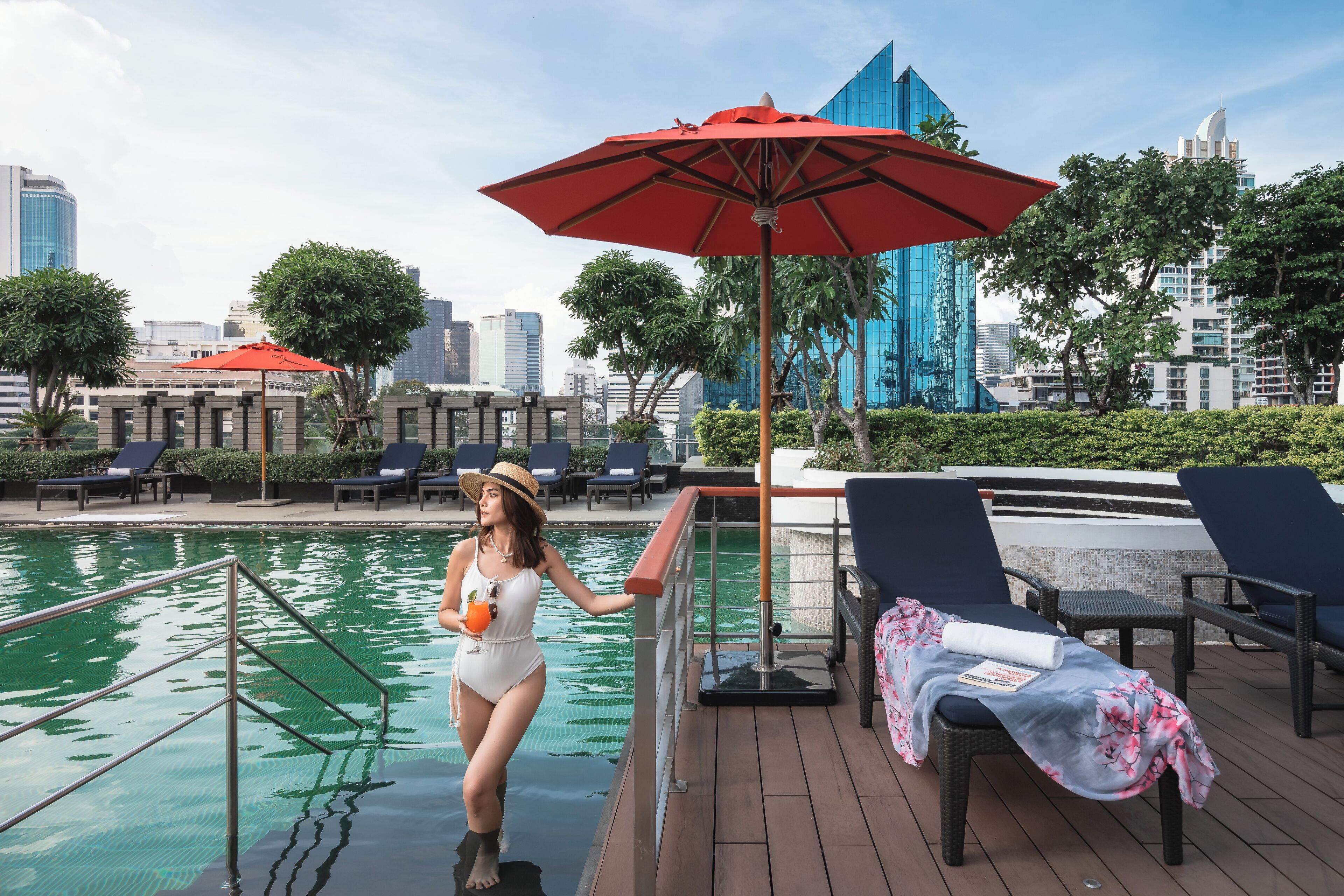 A poolside area with lounge chairs, a red umbrella, and a cityscape in the background.