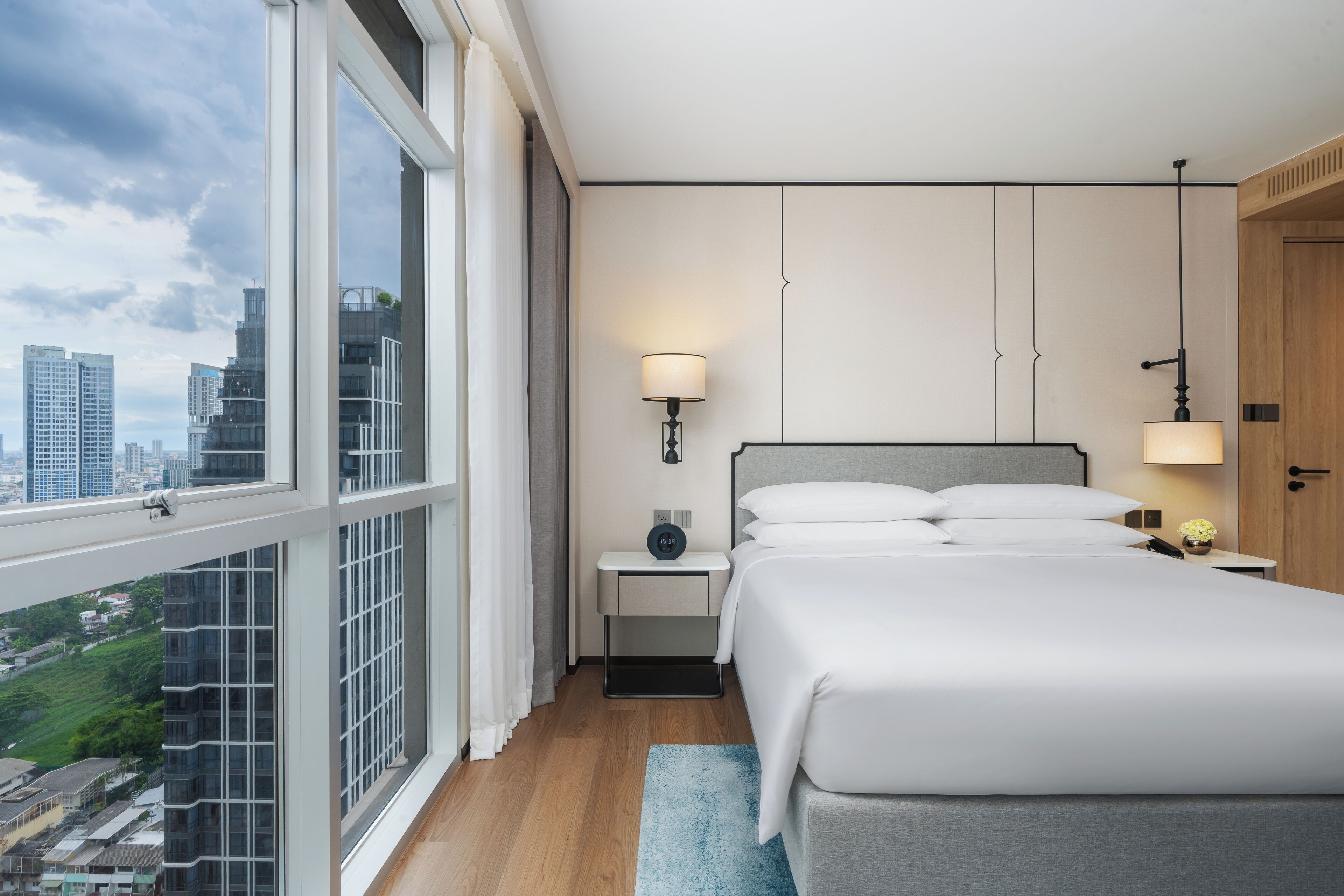 A modern hotel room with a large window, a bed with white linens, a bedside table with a lamp, and a view of the cityscape.