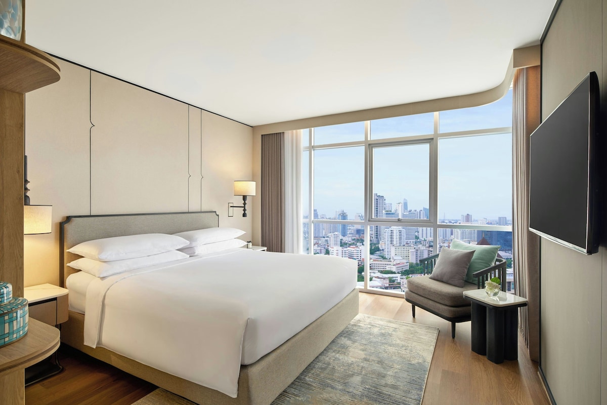 A modern hotel room with a large bed, a city view, a flat-screen TV, and a small seating area.