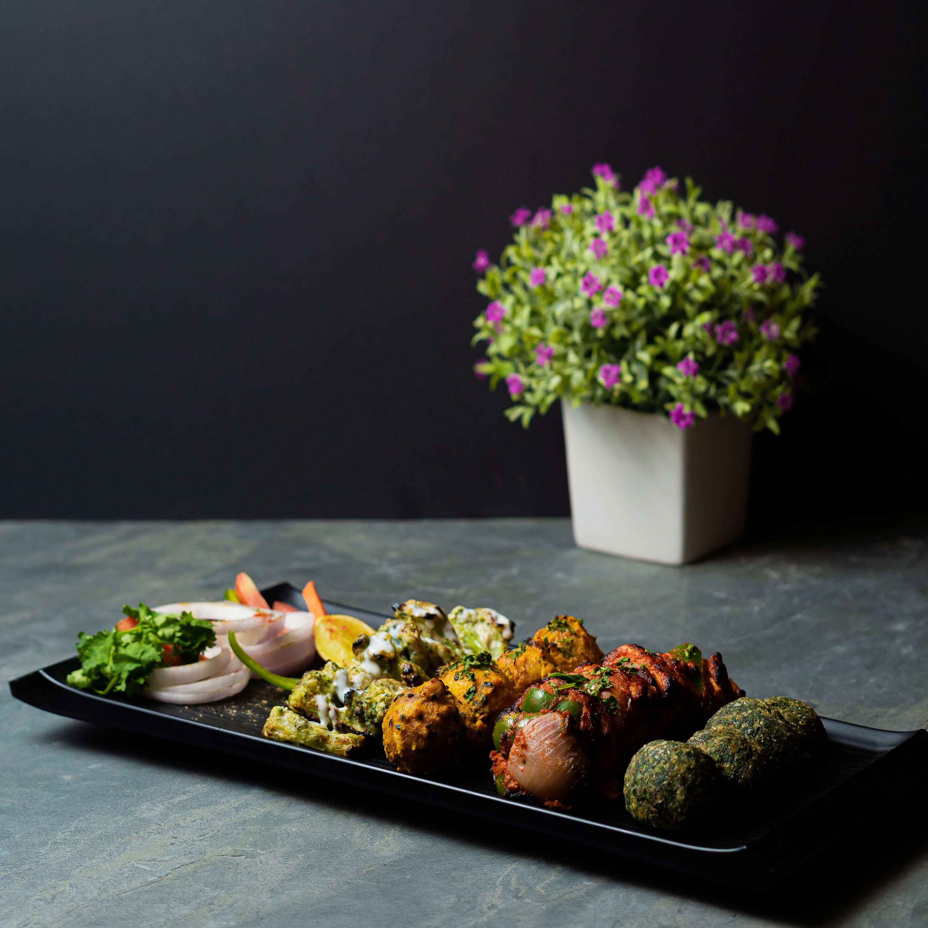 A plate of food with various dishes, including grilled meat and vegetables, on a dark surface with a potted plant in the background.