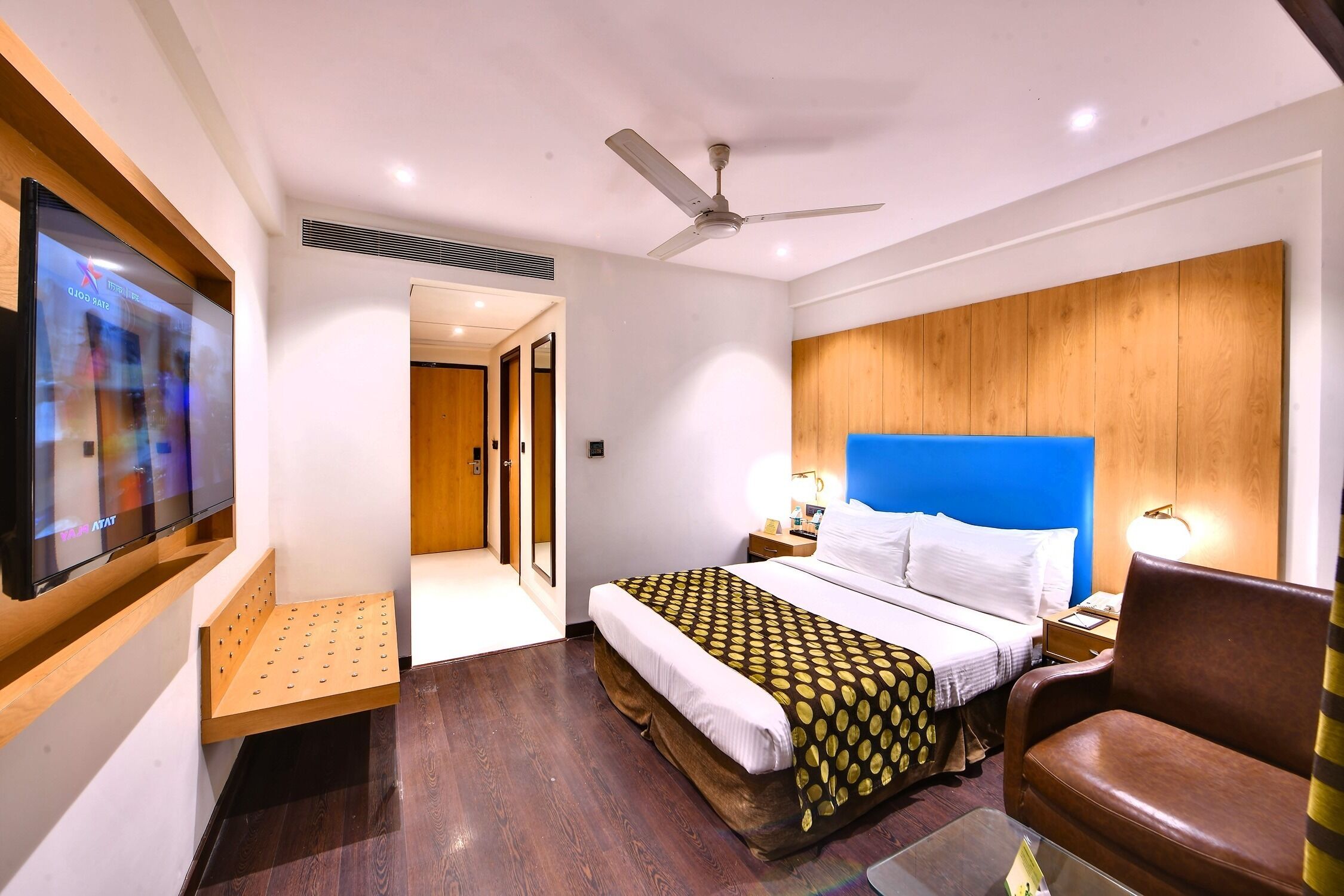 Habitación de hotel con cama, escritorio, silla, televisor y ventilador de techo.