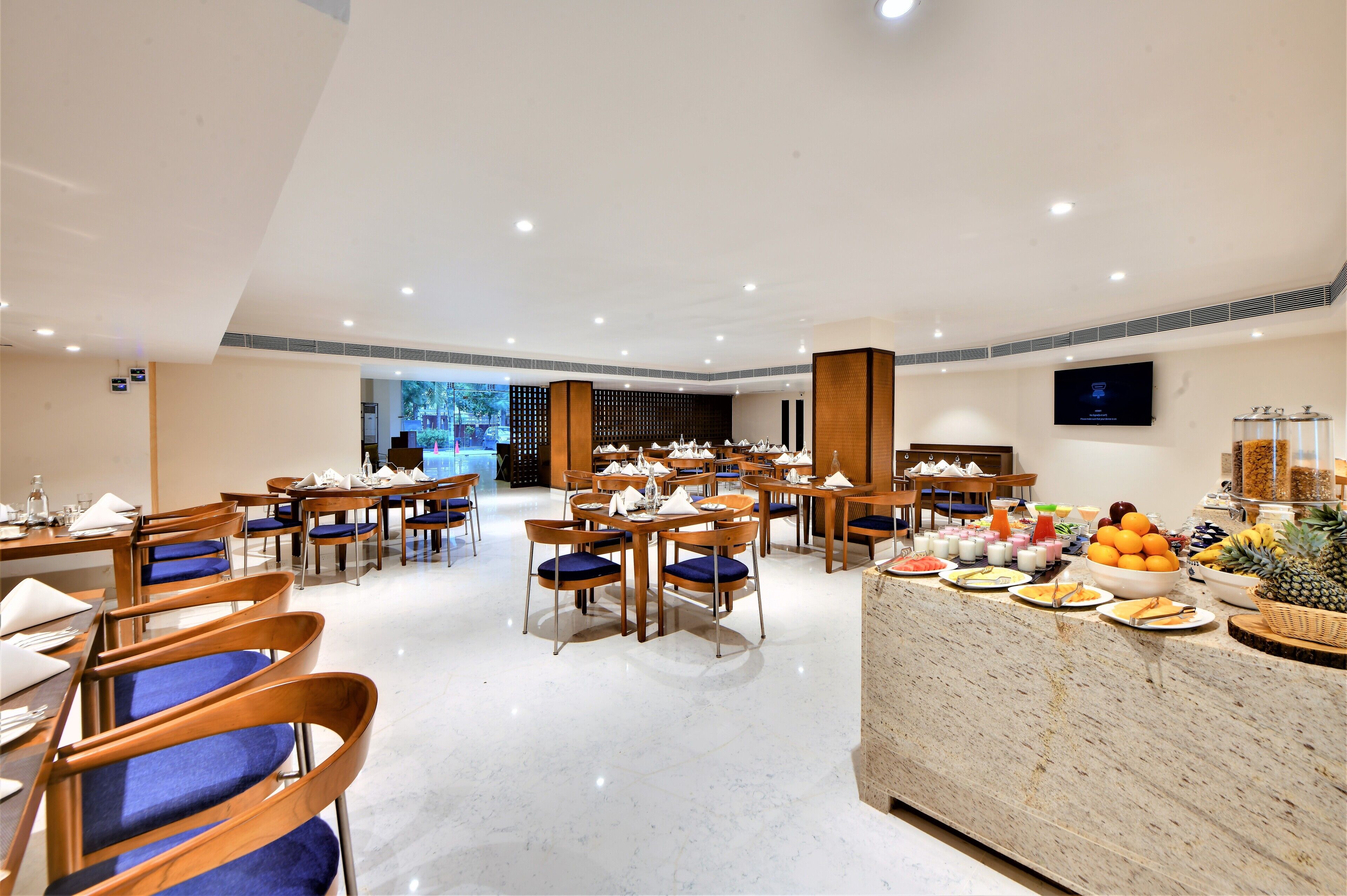 A spacious dining area with multiple tables and chairs, a buffet counter with various food items, and a large screen on the wall.