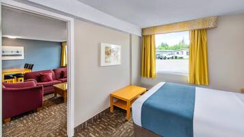 Habitación de hotel con cama, escritorio, sofá y una silla. Hay una ventana con cortinas y vista a un estacionamiento.