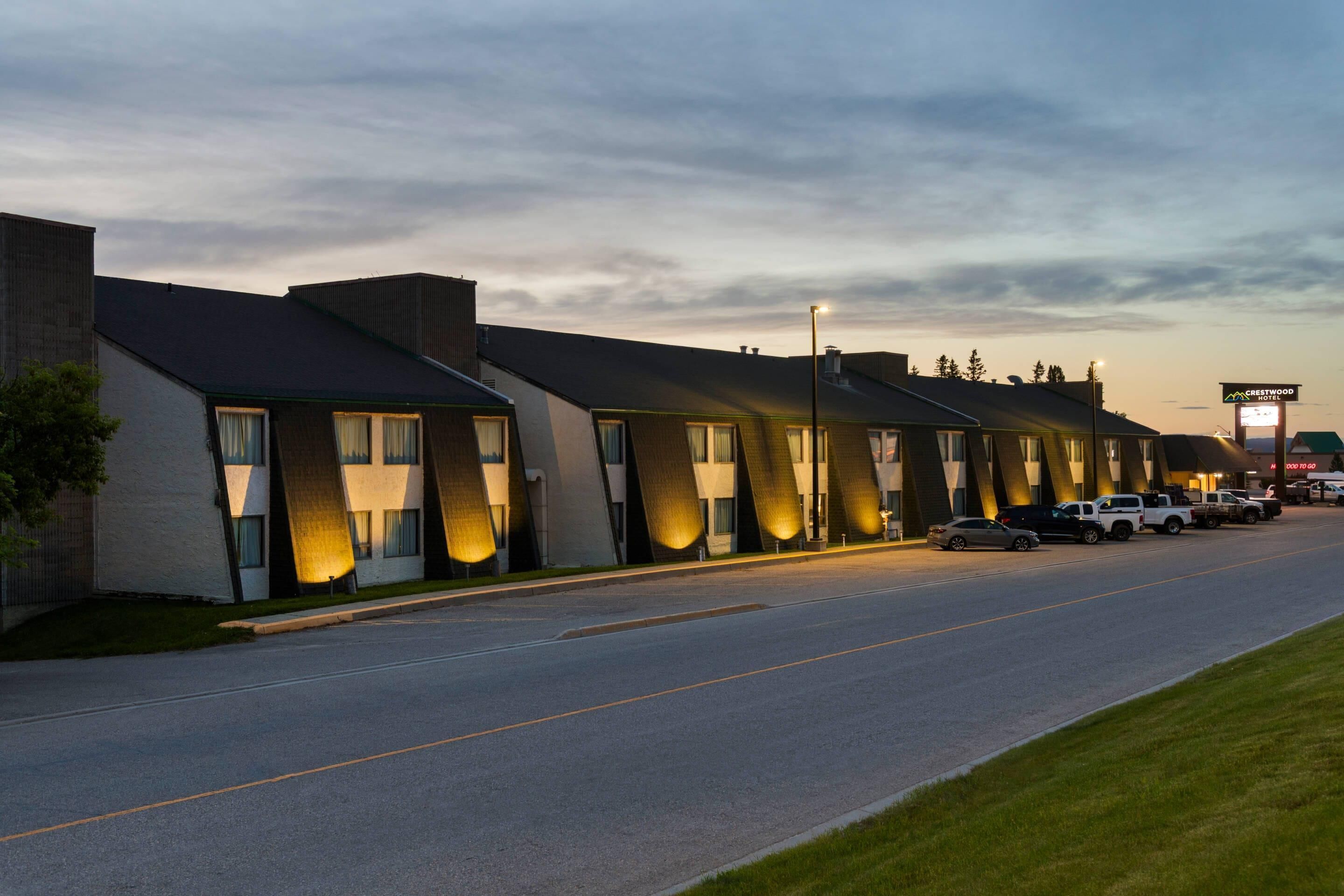 A row of modern motel buildings with illuminated windows, a clear sky, and a road with parked vehicles.