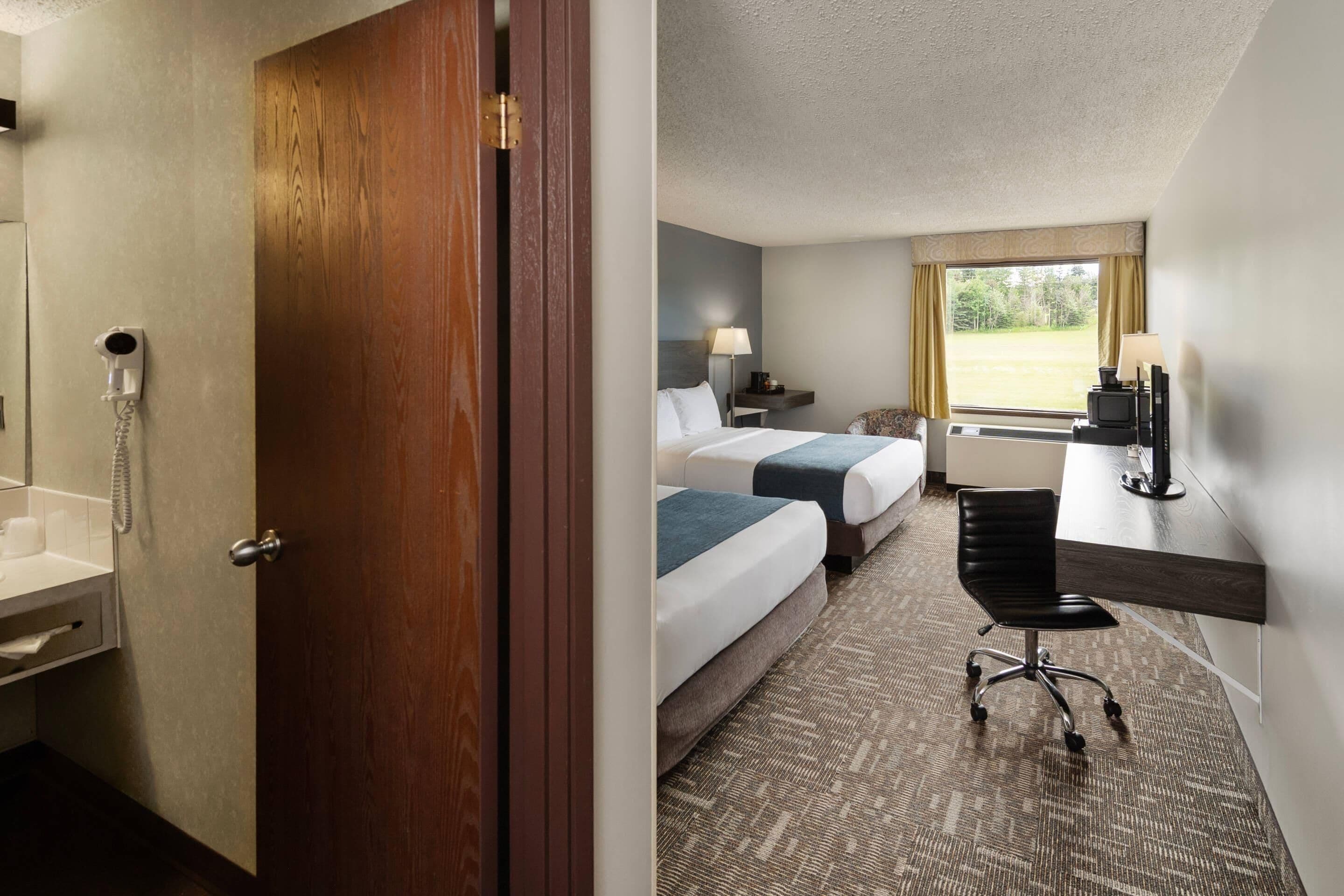 A hotel room with a wooden door, a bathroom with a telephone, two beds, a desk, a chair, and a window with a view.