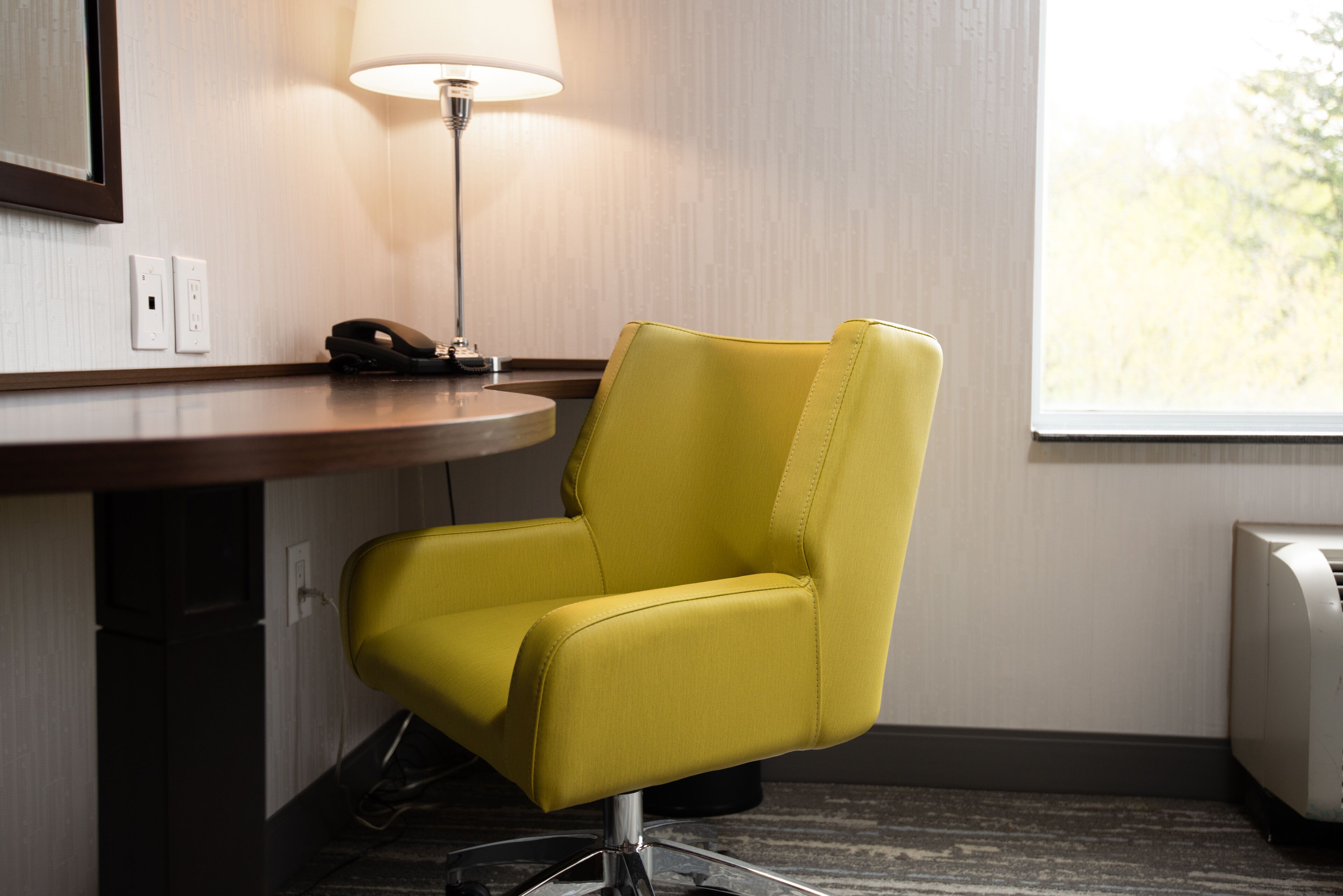 A yellow chair in a hotel room with a lamp, a phone, and a window.