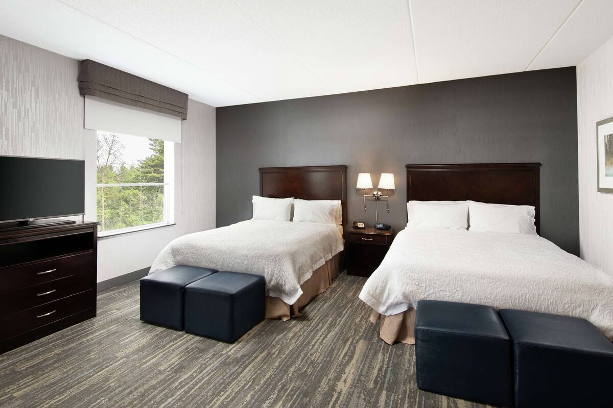 A hotel room with two beds, a TV, a desk, and a window with a view.