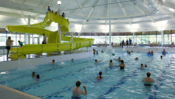 Uma grande piscina coberta com um escorregador amarelo e várias pessoas nadando e brincando.