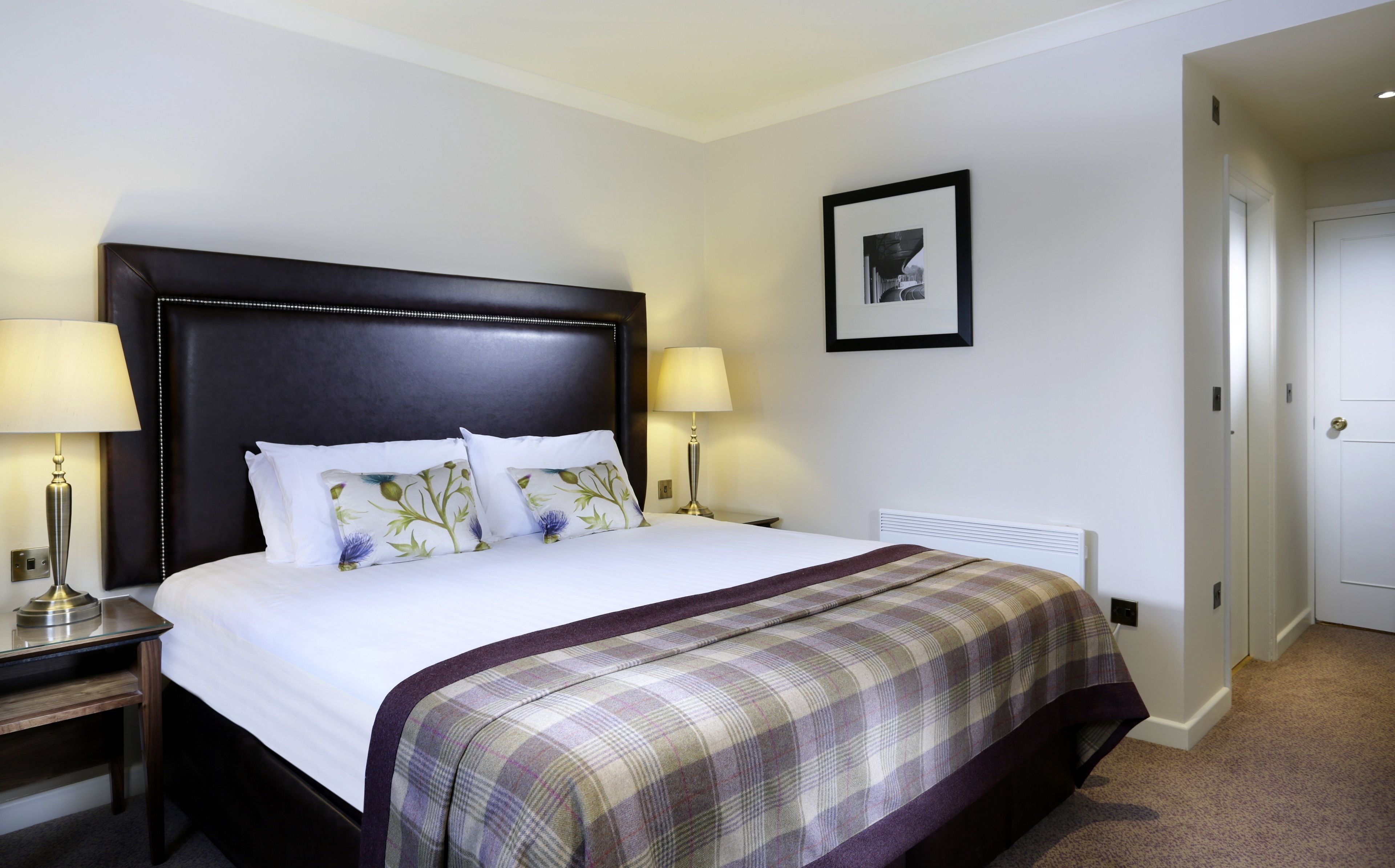 A hotel room with a large bed, two bedside lamps, a framed picture on the wall, and a door to the right.