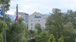 Pátio de hotel com área de estar externa, mastro com bandeira e edifício com letreiro 'avie more'.