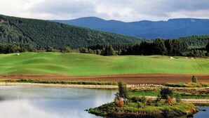 Um campo de golfe com um lago, colinas onduladas e uma cadeia montanhosa arborizada ao fundo.
