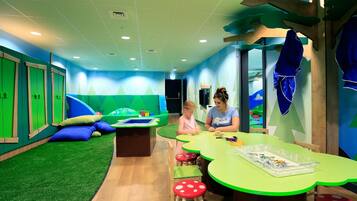 Zona de juegos infantiles con paredes verdes, una mesa de juegos y asientos coloridos.