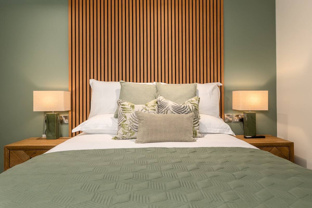 Una cama bien hecha con un edredón verde, dos lámparas de noche y un cabecero de listones de madera.