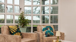 Una habitación con dos sillones color beige, un plaid verde, una mesa con lámpara y una ventana que ofrece vistas a árboles y edificios.