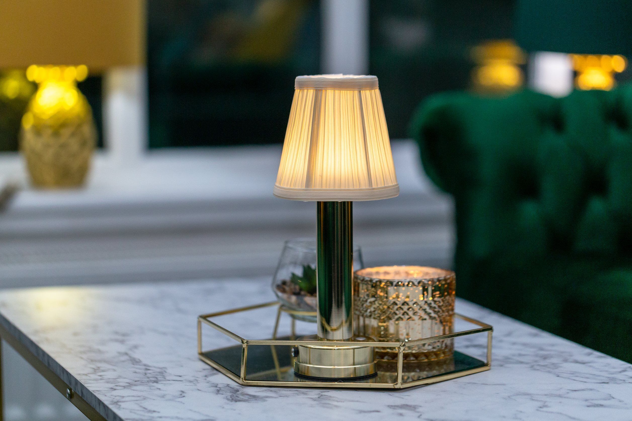 A table lamp with a pleated shade, a glass candle holder, and a gold-framed tray on a marble surface.