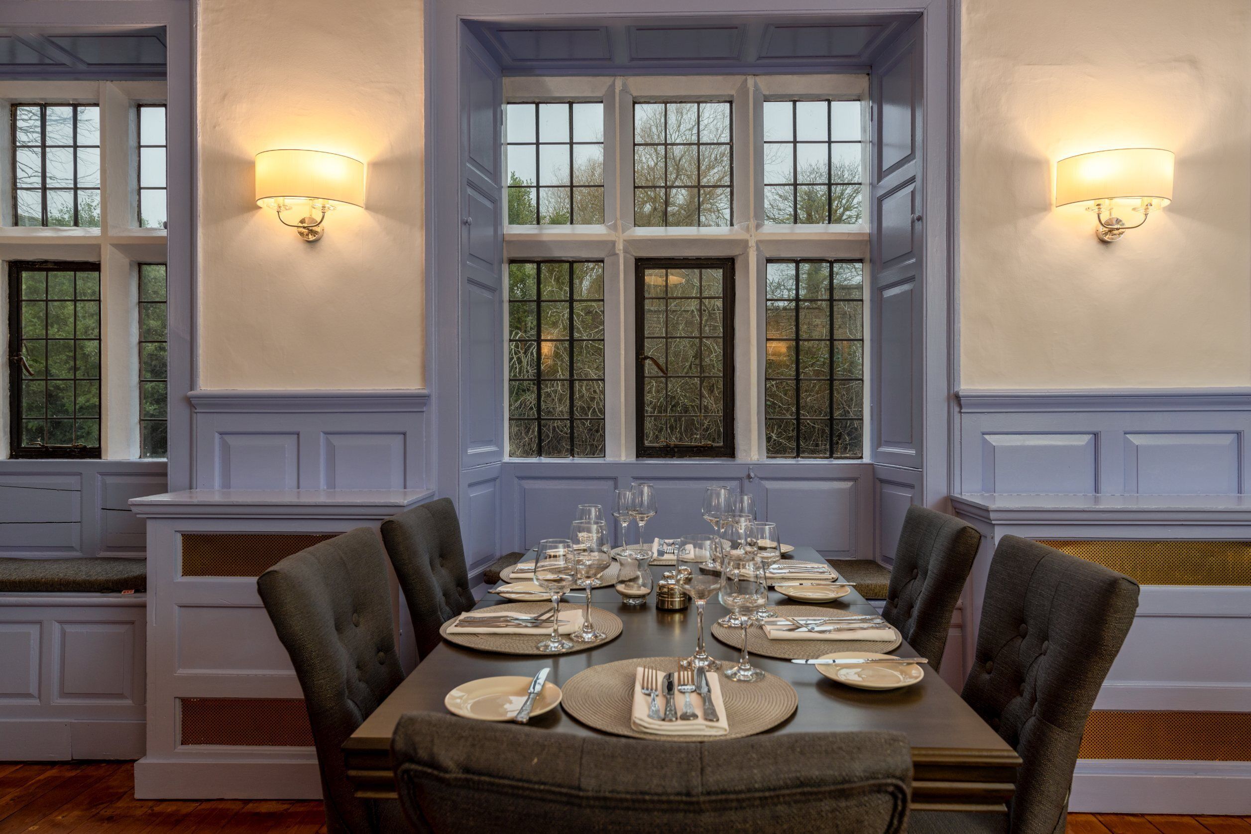Comedor con una mesa grande dispuesta para una comida, flanqueado por dos ventanas con lámparas.