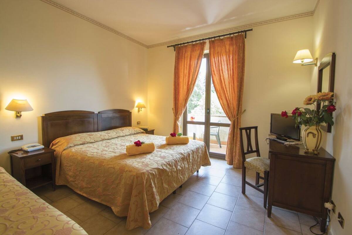 A hotel room with a large bed, two bedside tables with lamps, a desk with a television, a chair, and a window with curtains.