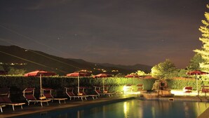 A nighttime pool area with lounge chairs and umbrellas, surrounded by a hedge and a mountain backdrop. - Park Hotel La Pineta (Mulazzo)