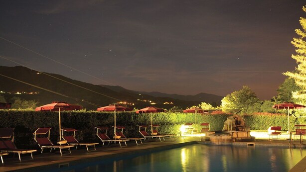 Zona de piscina nocturna con sillones de descanso y sombrillas, rodeada de setos y con montañas al fondo.