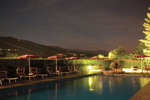 A nighttime pool area with lounge chairs and umbrellas, surrounded by a hedge and a mountain backdrop. - Park Hotel La Pineta (Mulazzo)