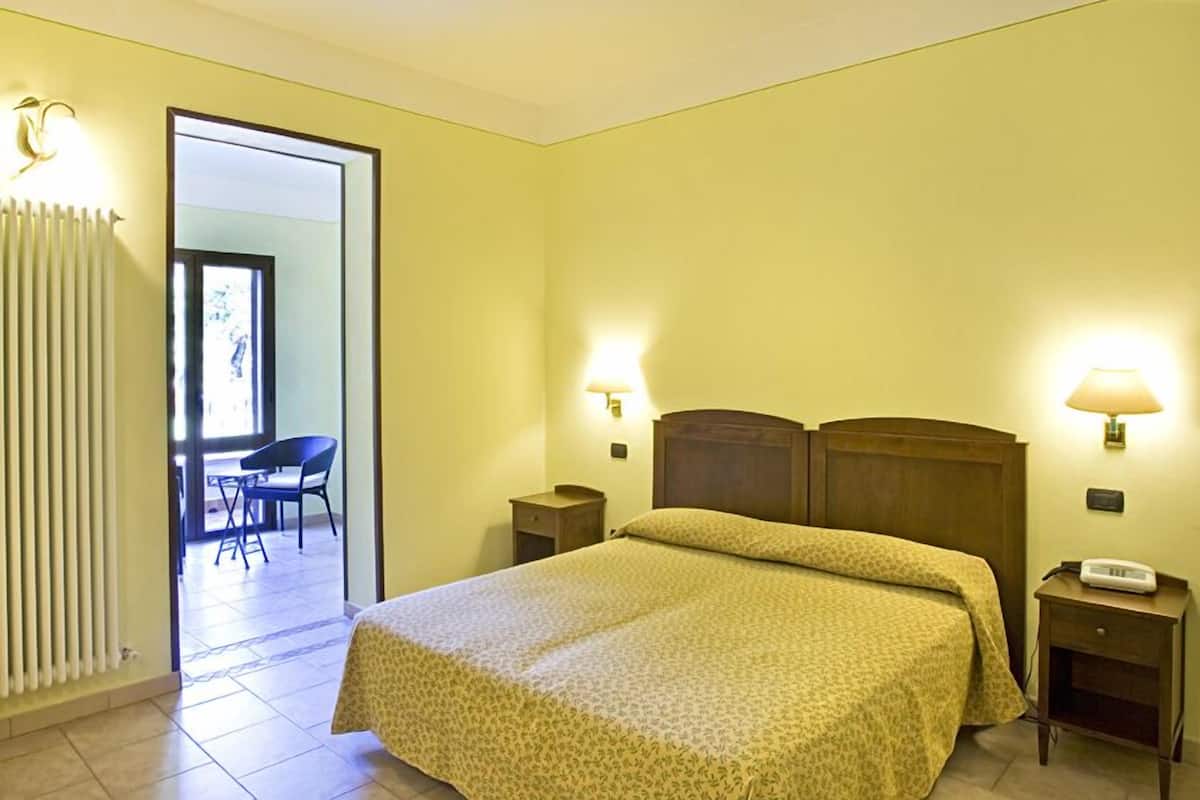 A hotel room with a bed, bedside lamps, a nightstand, and a chair in the adjacent room.