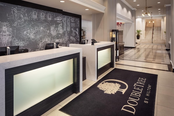Zona de recepción de hotel con diseño moderno, que incluye un mostrador de recepción, una pizarra con dibujos técnicos y una alfombra con el nombre del hotel.