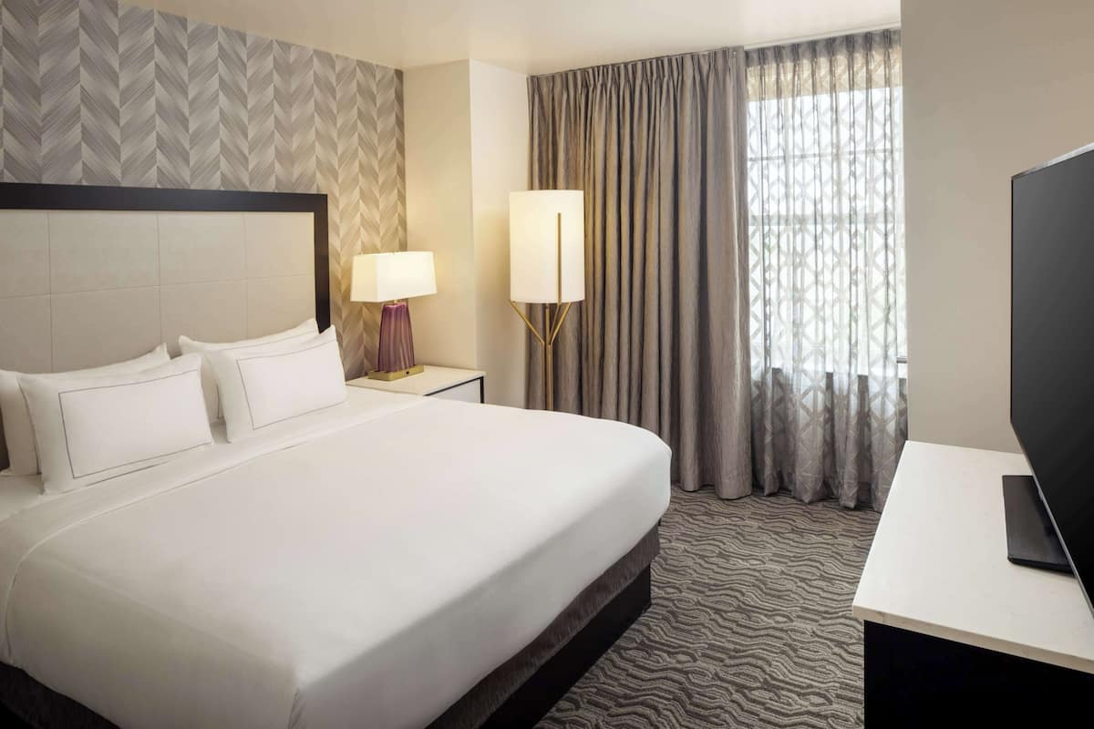 A hotel room with a large bed, a bedside table with a lamp, a TV, and a window with curtains.