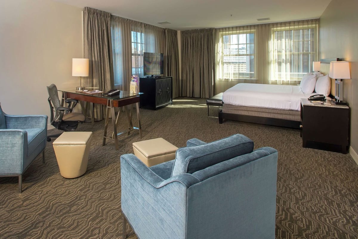 A hotel room with a bed, two armchairs, a desk with a computer, a television, and a window with curtains.