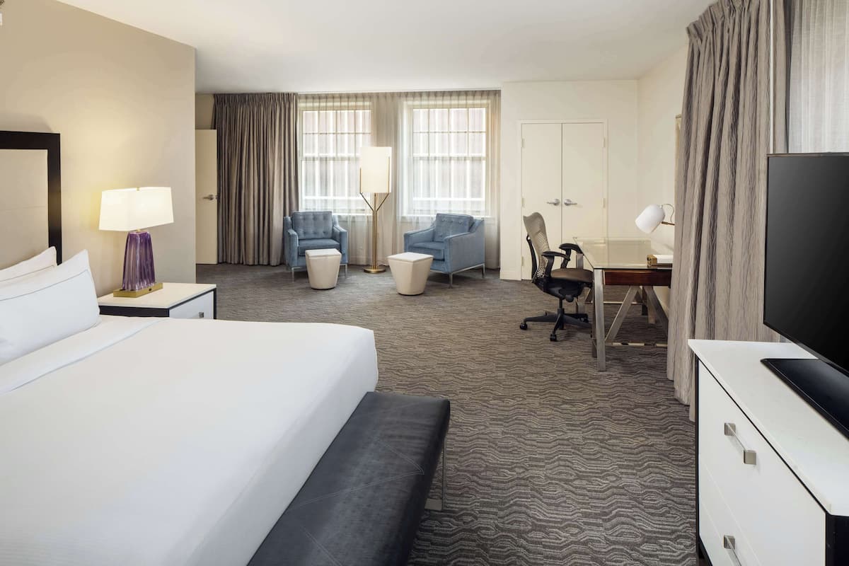 A hotel room with a large bed, two armchairs, a desk with a chair, a television, and a wardrobe.