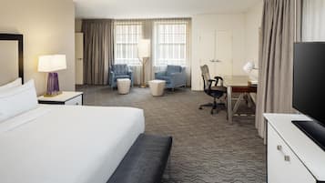 Quarto de hotel com cama, escrivaninha, cadeira, televisão e janela com cortinas.
