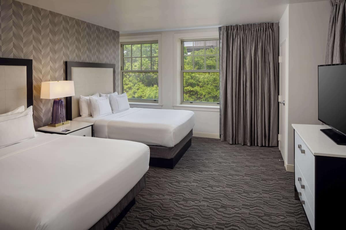 A hotel room with two beds, a TV, a lamp, and a window with a view of greenery.