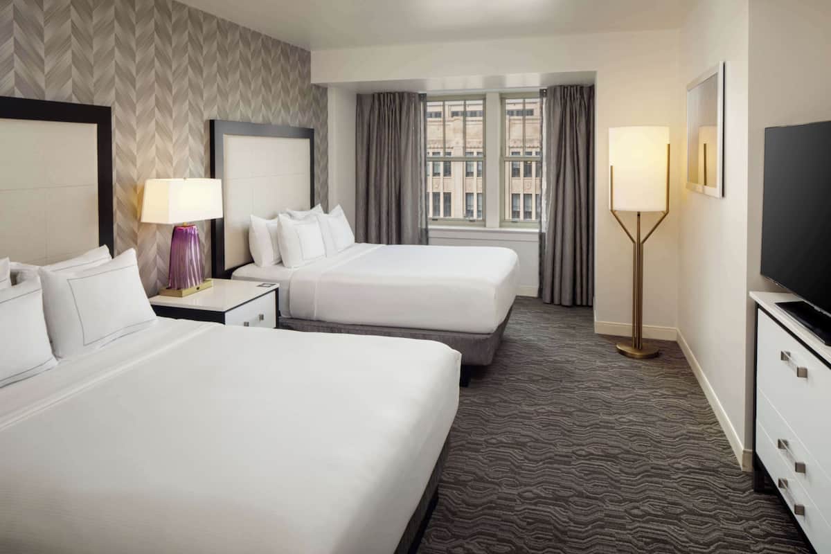 A hotel room with two beds, a large window with curtains, a floor lamp, and a TV on a dresser.