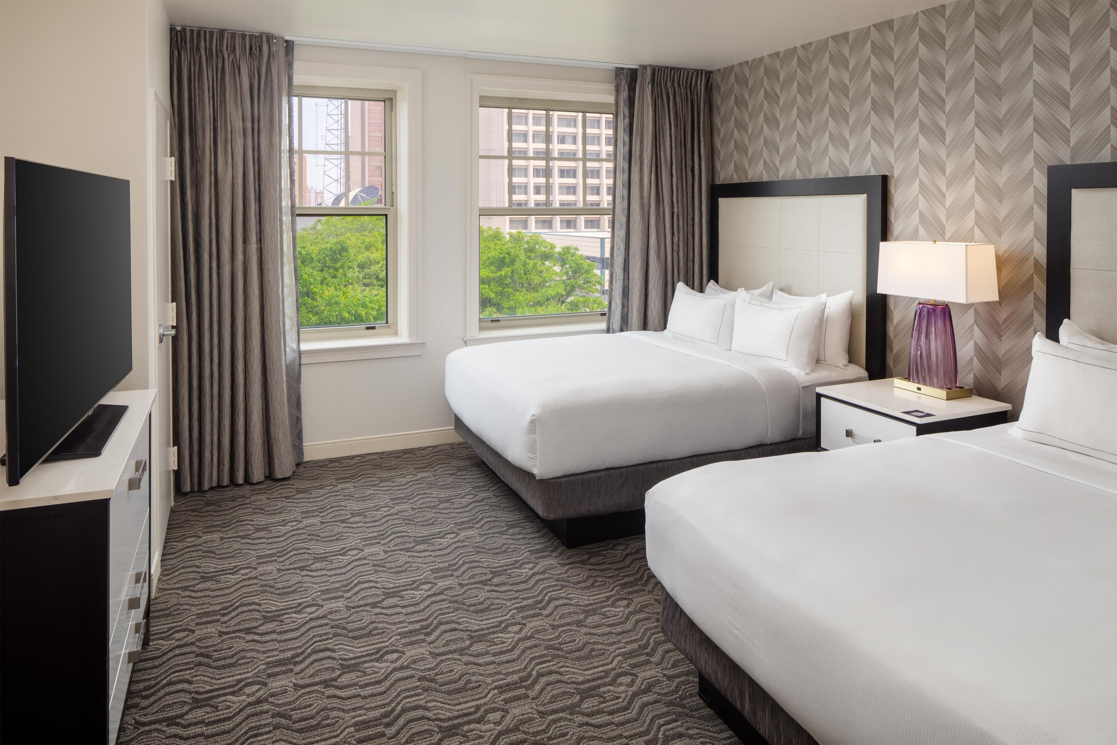A hotel room with two beds, a TV, a nightstand with a lamp, and a view of the city through the windows.