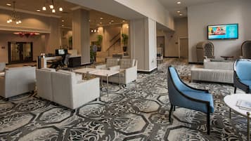 Un hall d’entrée spacieux, doté d’un tapis à motifs, de plusieurs espaces de détente comprenant des canapés et des fauteuils, d’un escalier et d’un téléviseur fixé au mur.