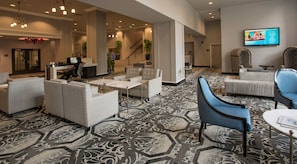 A spacious lobby with a patterned carpet, multiple seating areas including sofas and armchairs, a staircase, and a television mounted on the wall. - DoubleTree Suites by Hilton Hotel Detroit Downtown - Fort Shelby (Detroit)
