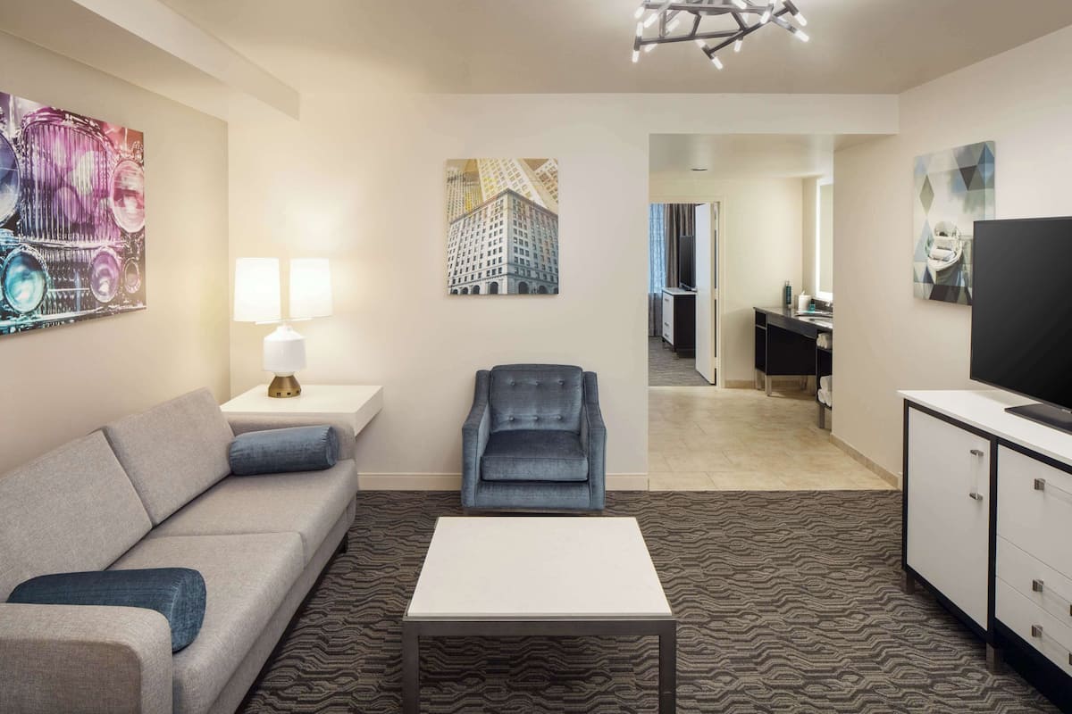 A hotel room with a sofa, armchair, TV, and artwork on the walls.