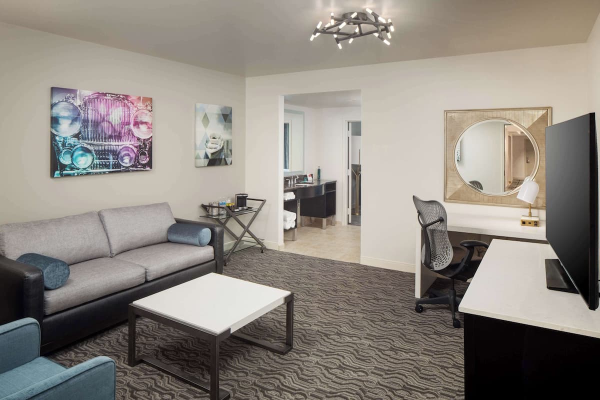 A modern living room with a sofa, coffee table, and TV. There is a dining area with a table and chairs. The room has artwork on the walls and a large mirror.