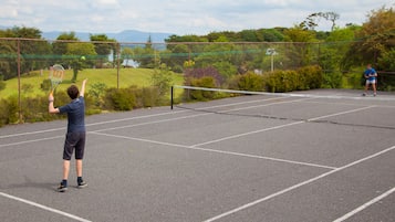 A tennis court with a net, surrounded by a chain-link fence and greenery.