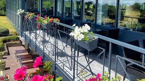 Un espacio exterior con mesas y sillas de metal, rodeado de grandes ventanales y plantas en macetas.