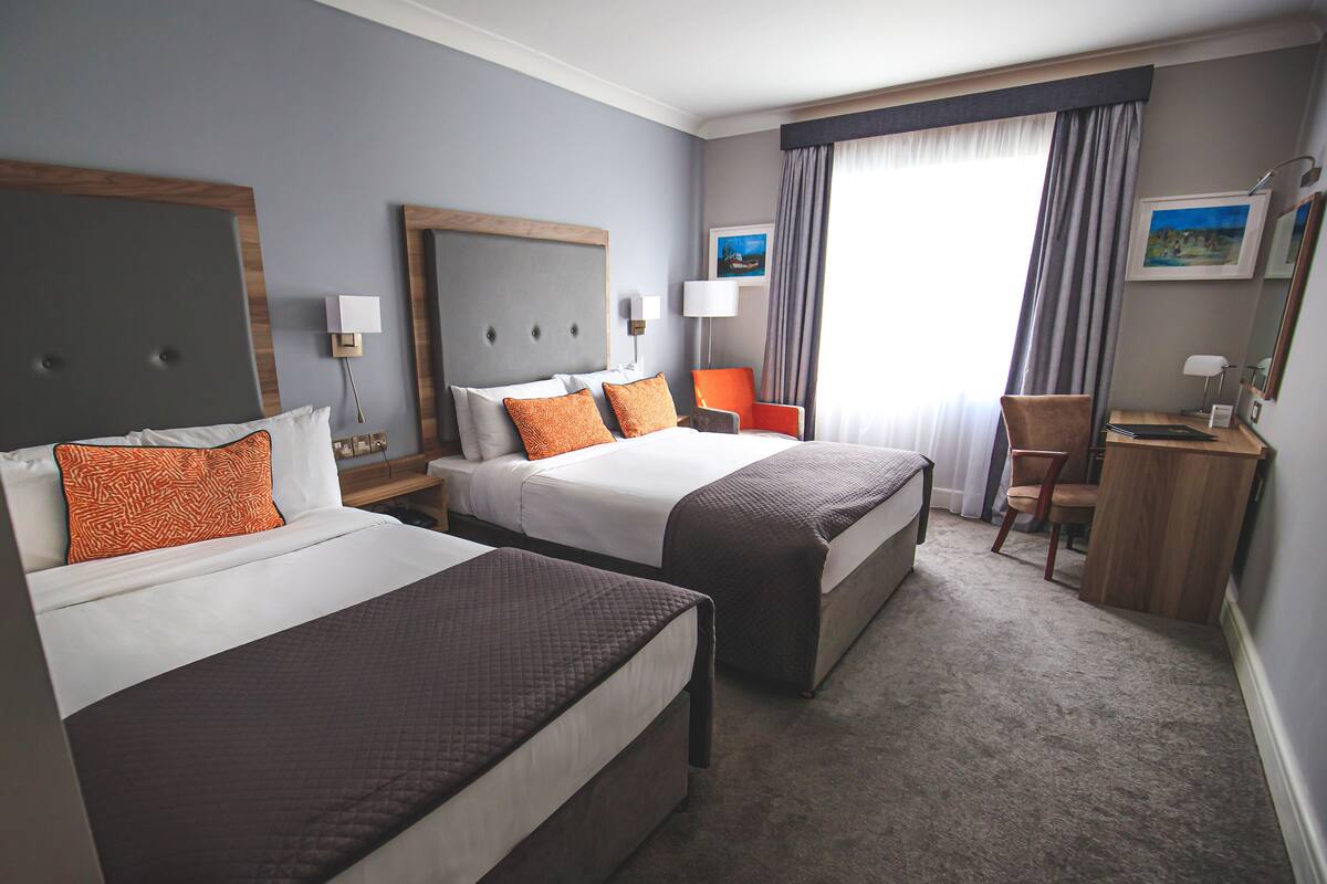 A hotel room with two beds, a desk, a chair, and a large window with curtains.