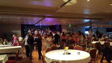A crowded indoor event with round tables and chairs, a stage in the background, and a ceiling with recessed lighting.