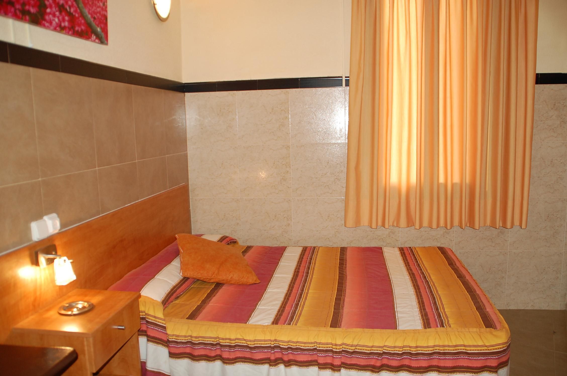 Una habitación de hotel con cama, mesita de noche, lámpara y ventana con cortinas.