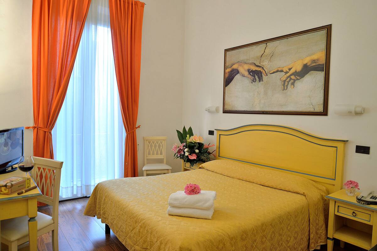 A hotel room with a bed, a desk with a computer, a chair, and a nightstand with a phone and a vase of flowers.