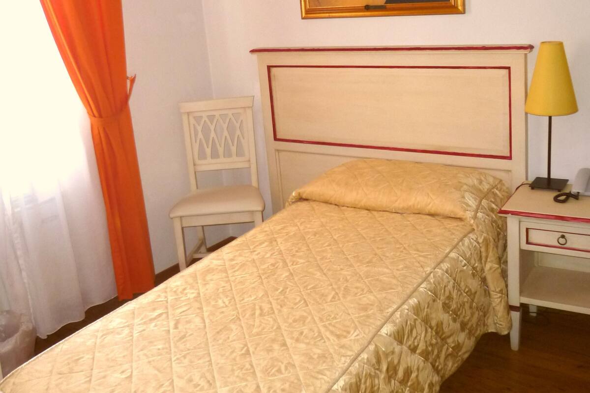 A single bed with a quilted cover, a bedside table with a lamp, a chair, and a framed picture on the wall.