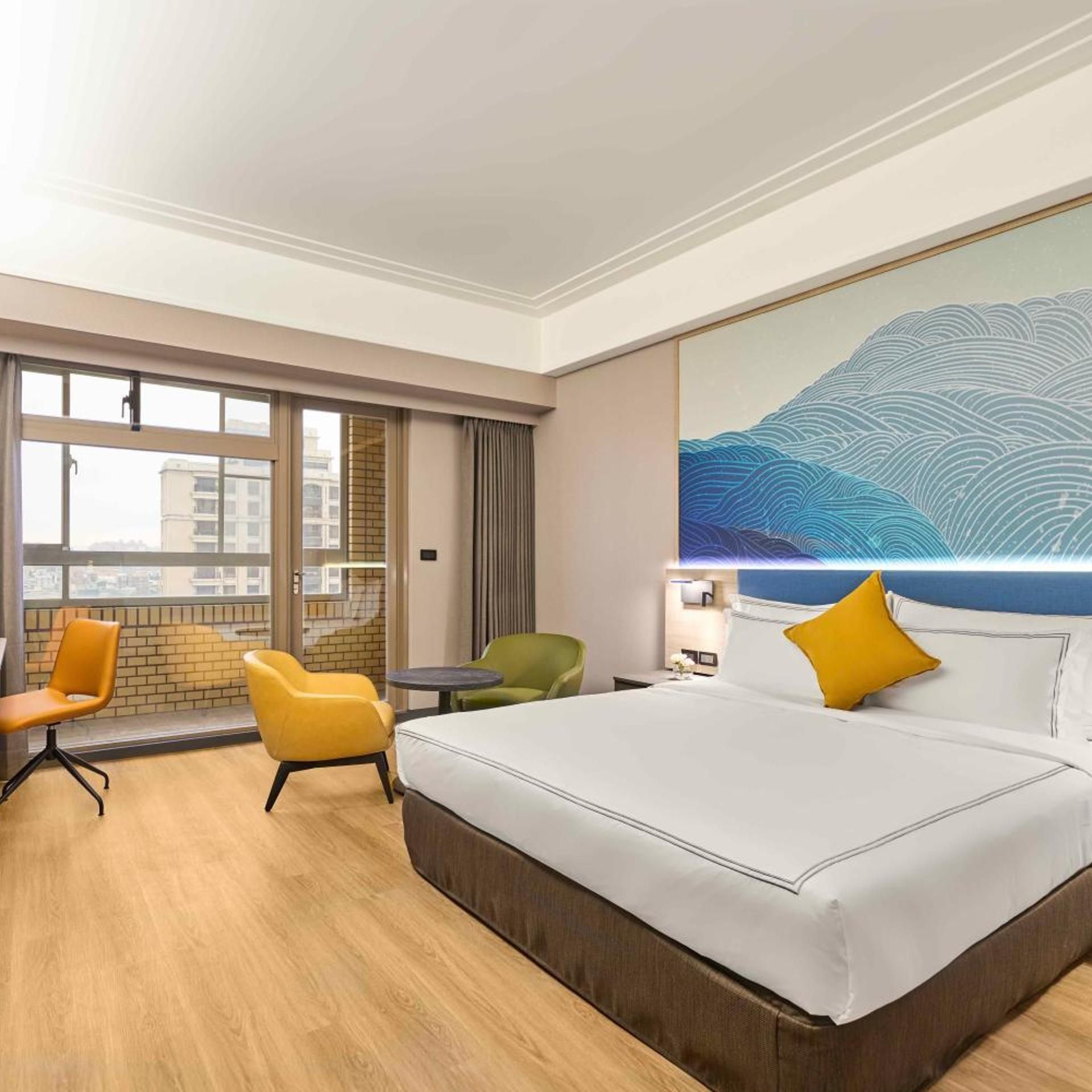 Une chambre d’hôtel avec un grand lit, deux fauteuils, une petite table et une vue sur la ville grâce à de grandes fenêtres.