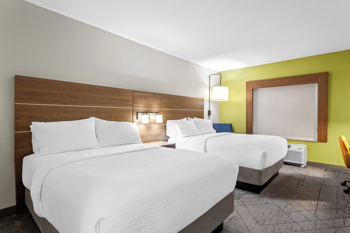 A hotel room with two beds, a nightstand, a lamp, a window, and a chair.